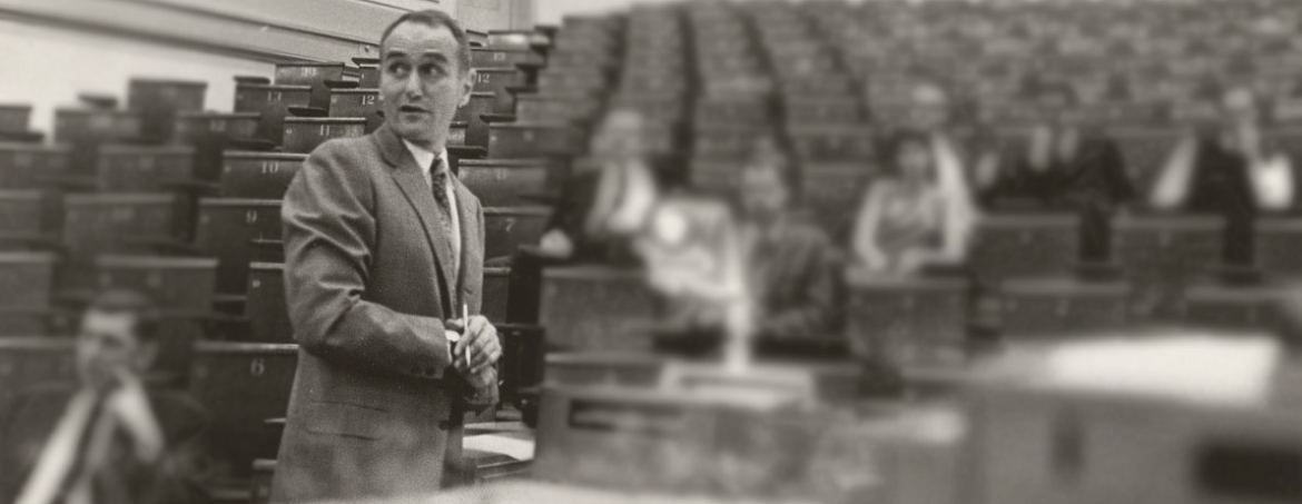 Professor Daniel M. Stressguth (1924-2020) in an old black and white photo standing at the front of a lecture hall with blurred people in the background