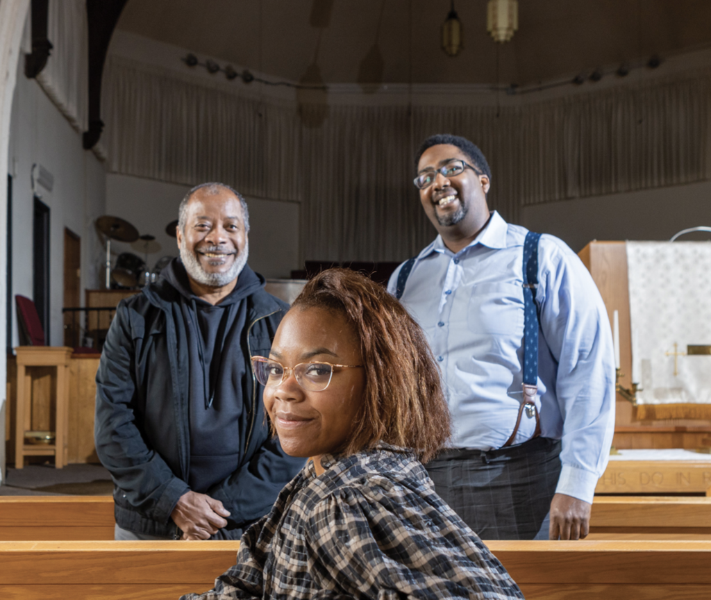 Three people smiling, where two men are standing and one woman is sitting in a church pew