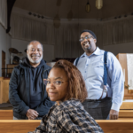 Three people smiling, where two men are standing and one woman is sitting in a church pew