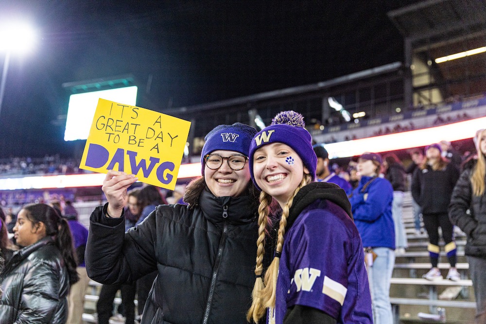 Two students at a football game holding a sign that says "It's a great day to be a dawg"