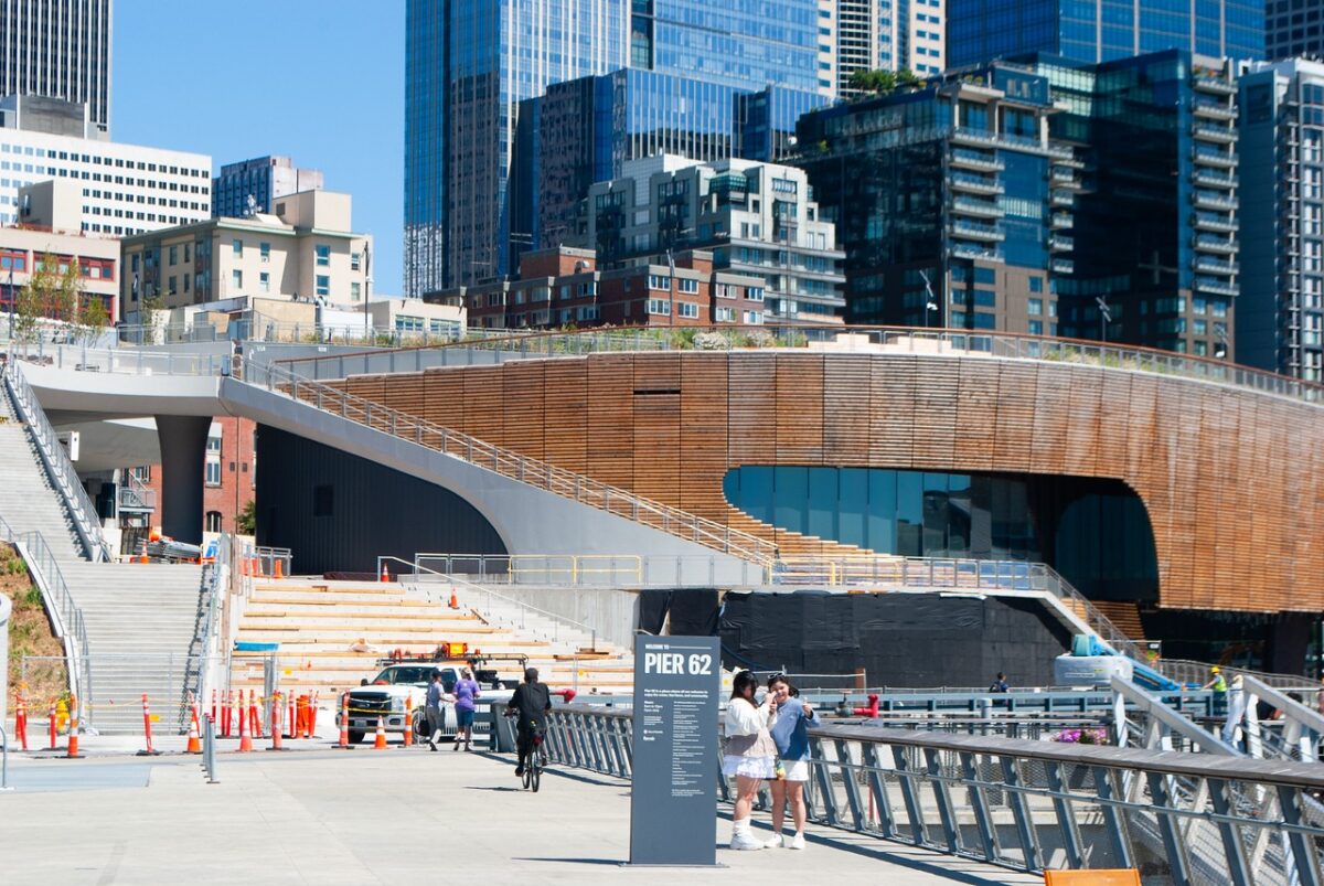 People walking on a pier with a sign that says "Pier 62" in front of an intricate wooden building