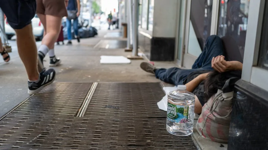 A person lying on a city sidewalk with a water bottle beside them as pedestrians pass by.