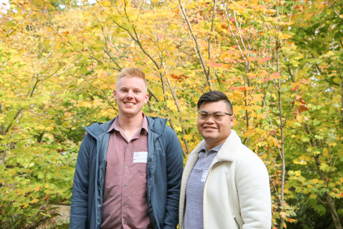 Two people standing together smiling in front of some trees