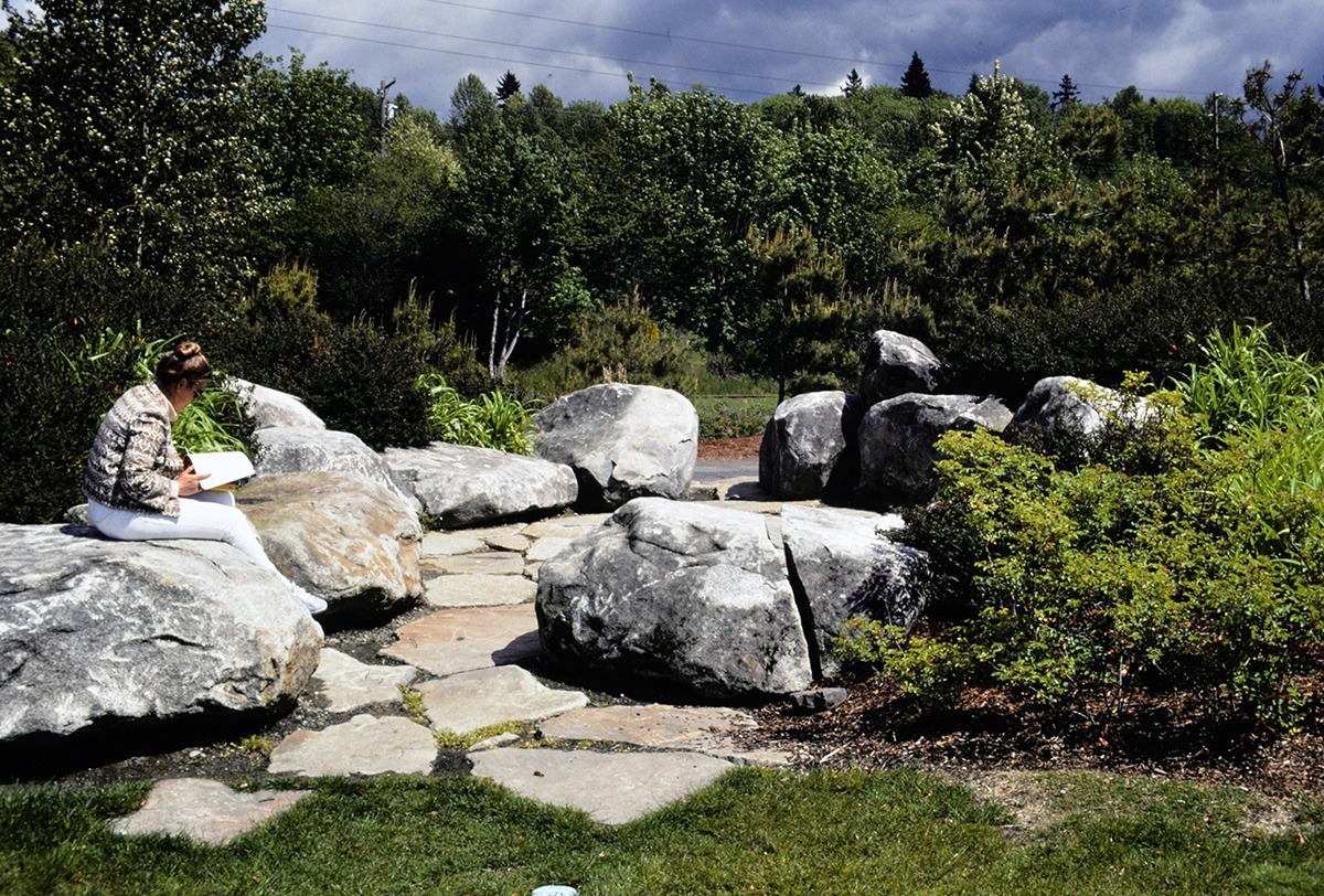 A person sitting on a large rock in a park reading a book