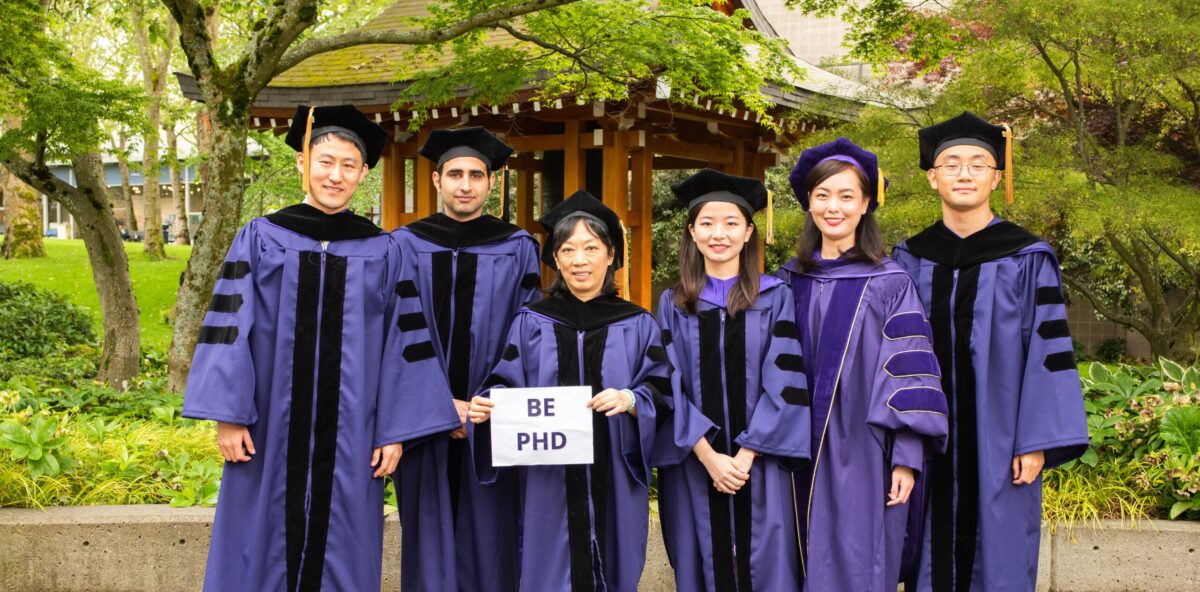 Group of students in purple robes holding up sign that says, "BE PHD"