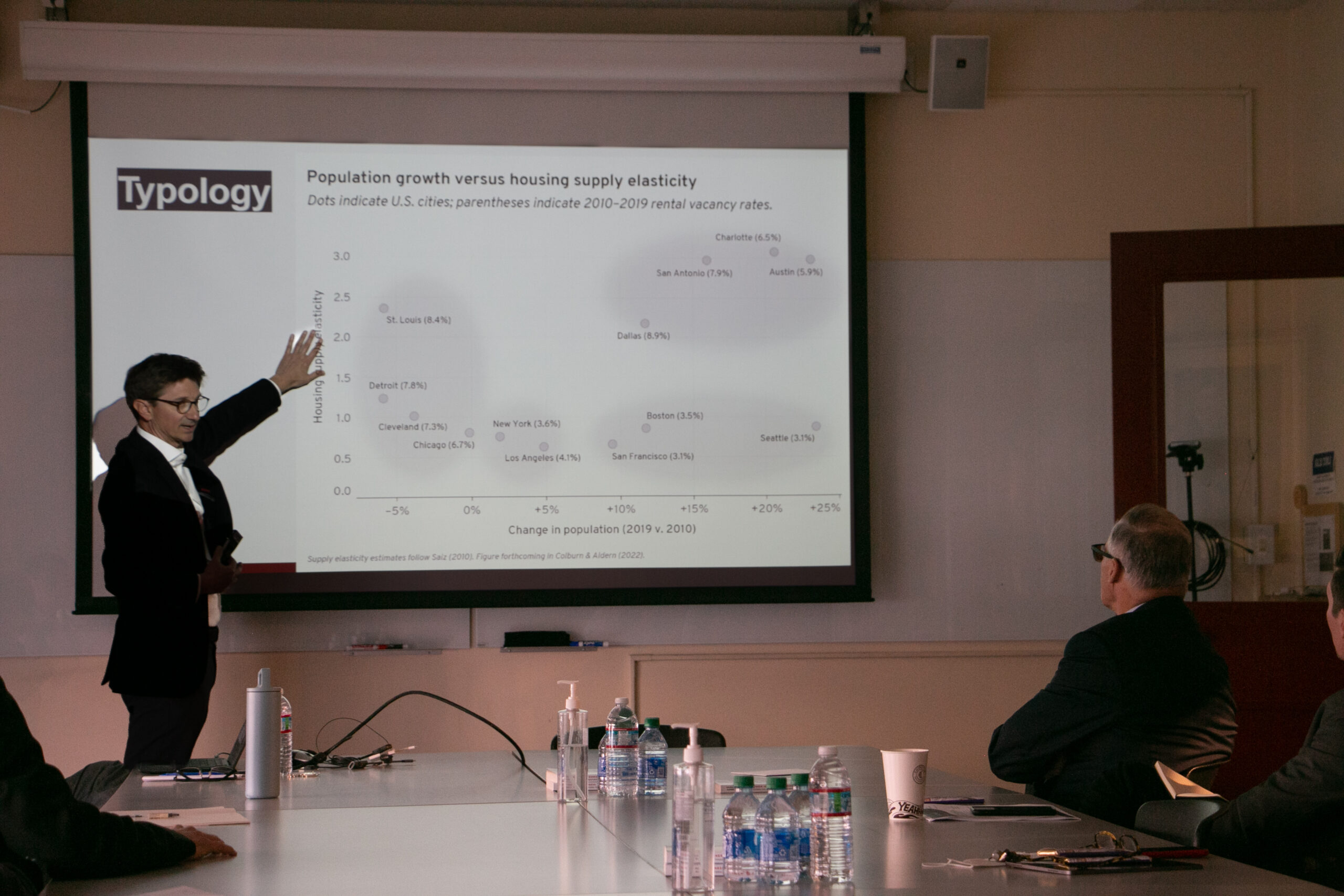Professor presenting in front of a screen to a man sitting at a table
