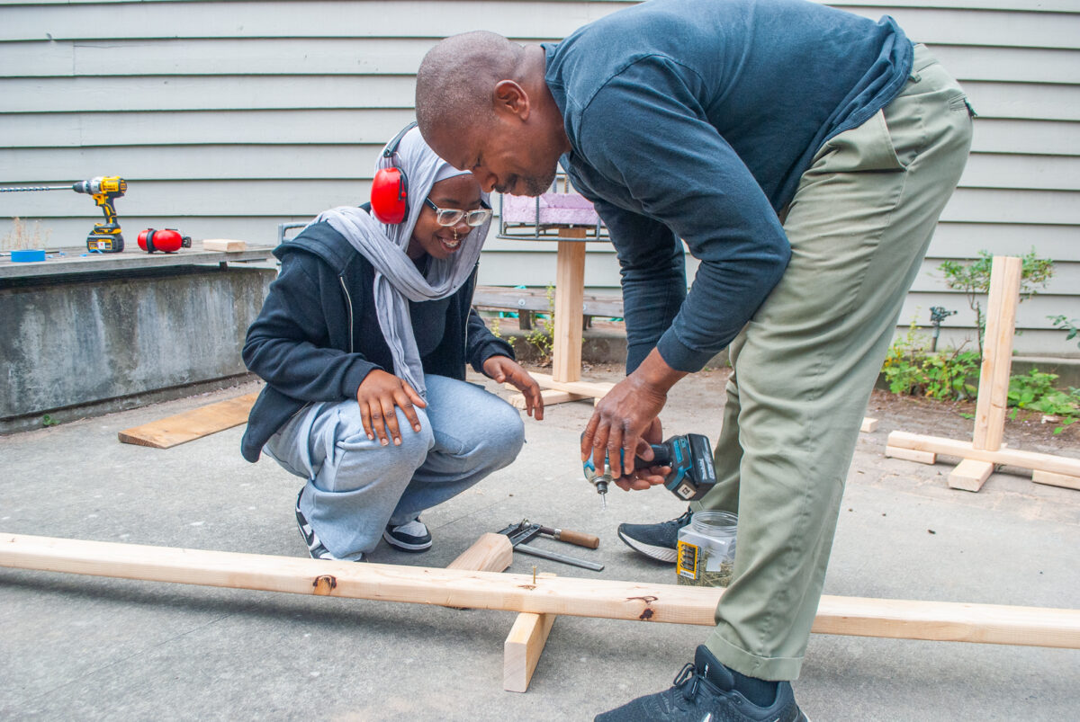 A person teaching a young person how to drilling a screw into two pieces of wood