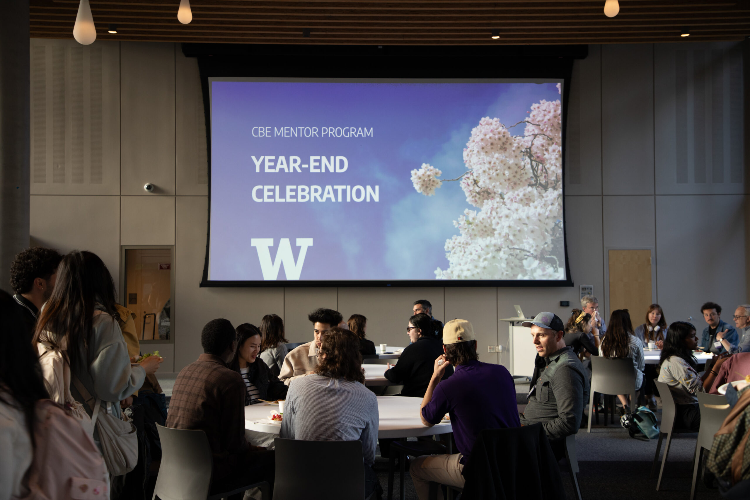 Students, mentors, and faculty gather in a sunlit, modern event space for the College of Built Environments Mentorship Program Year-End Celebration. A large screen at the front displays a purple UW slide with cherry blossoms and the text “CBE Mentor Program Year-End Celebration.” Tables are full, and a speaker addresses the audience from a podium.