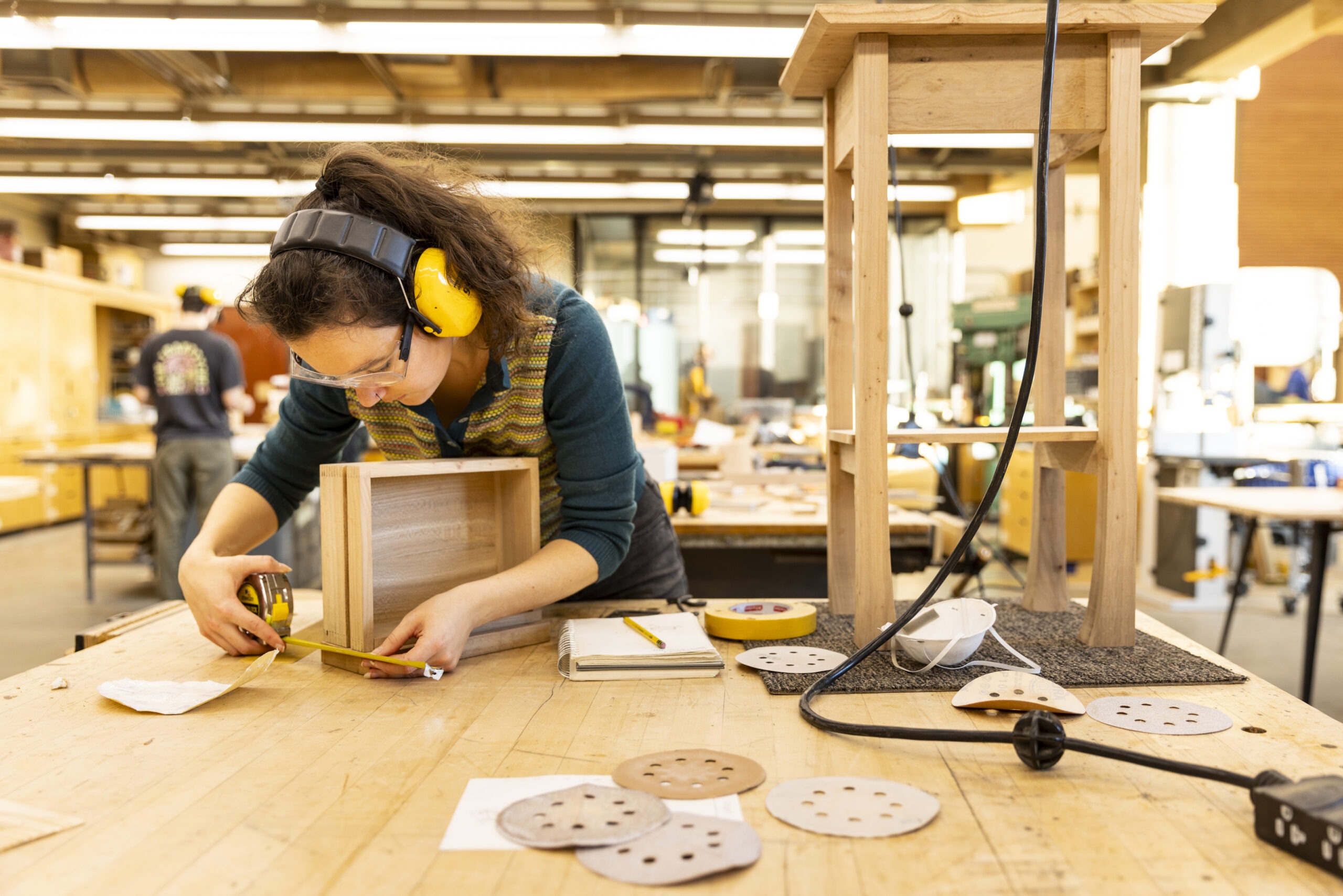 A student in safety goggles and yellow ear protection carefully measures a small wooden drawer on a workbench filled with tools, sandpaper disks, and a wooden side table in progress.