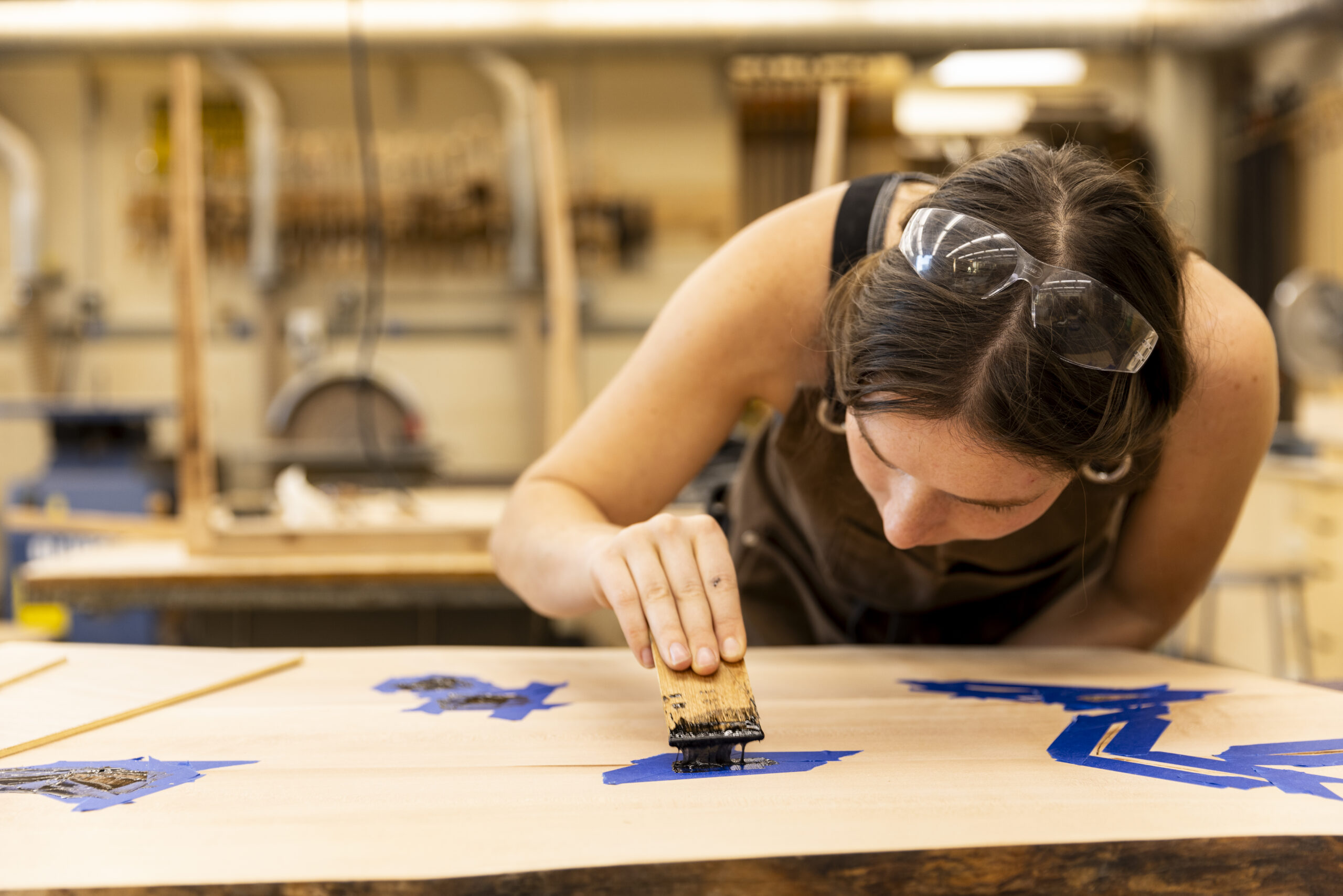 A student applies black paint or resin to a masked-off section of a wooden panel using a brush, concentrating on detail work while wearing safety goggles and an apron.