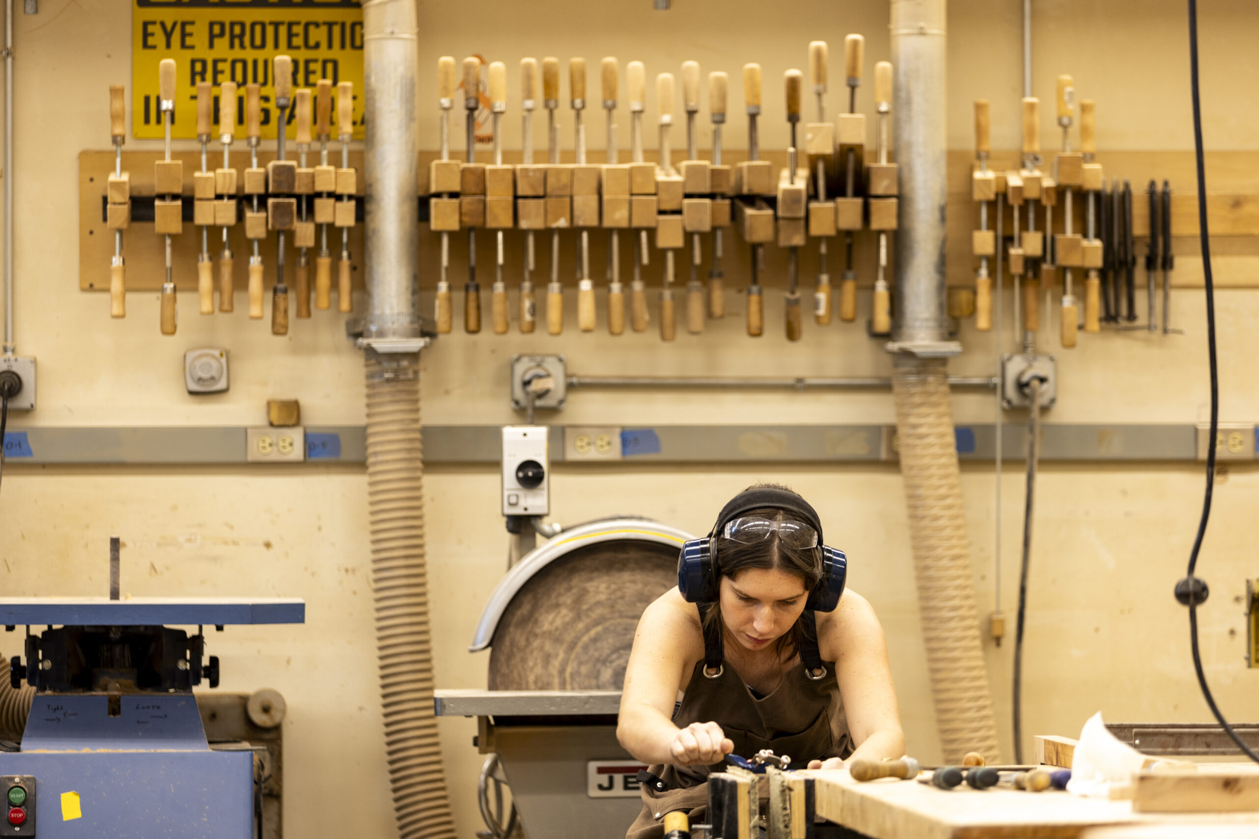 A student wearing hearing protection and a brown apron works intently on a piece of wood using a hand tool, with a wall of woodworking chisels and tools behind her.