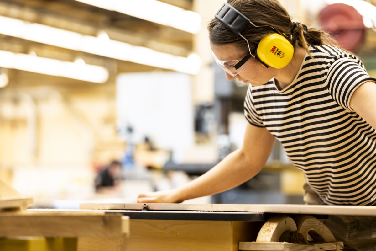 A student in a striped shirt uses a table saw to cut a piece of wood, pushing the material forward while wearing hearing protection and safety glasses in a bright, busy workshop.