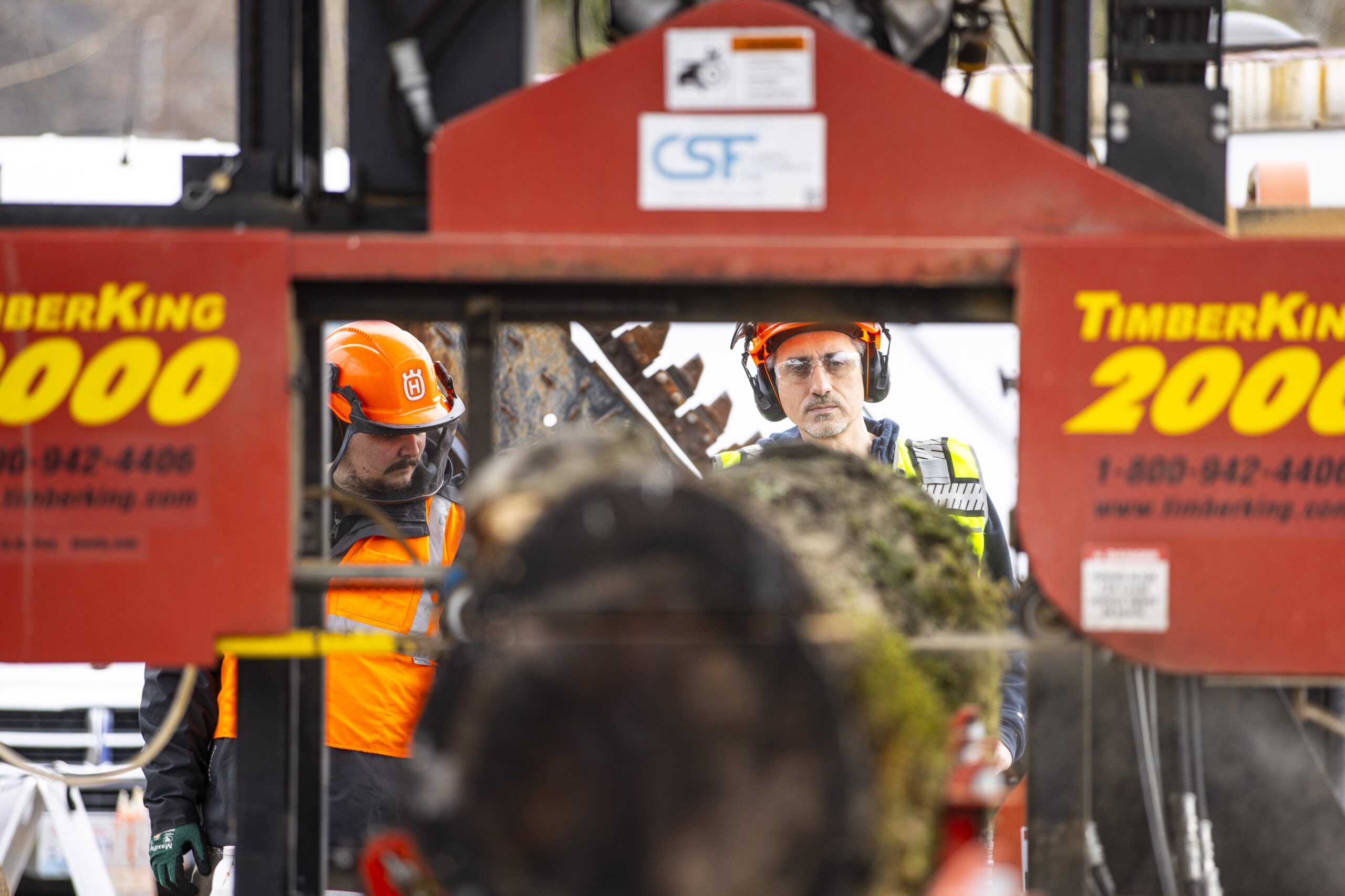 Two workers in orange safety vests and helmets monitor a log being processed through a TimberKing 2000 sawmill.