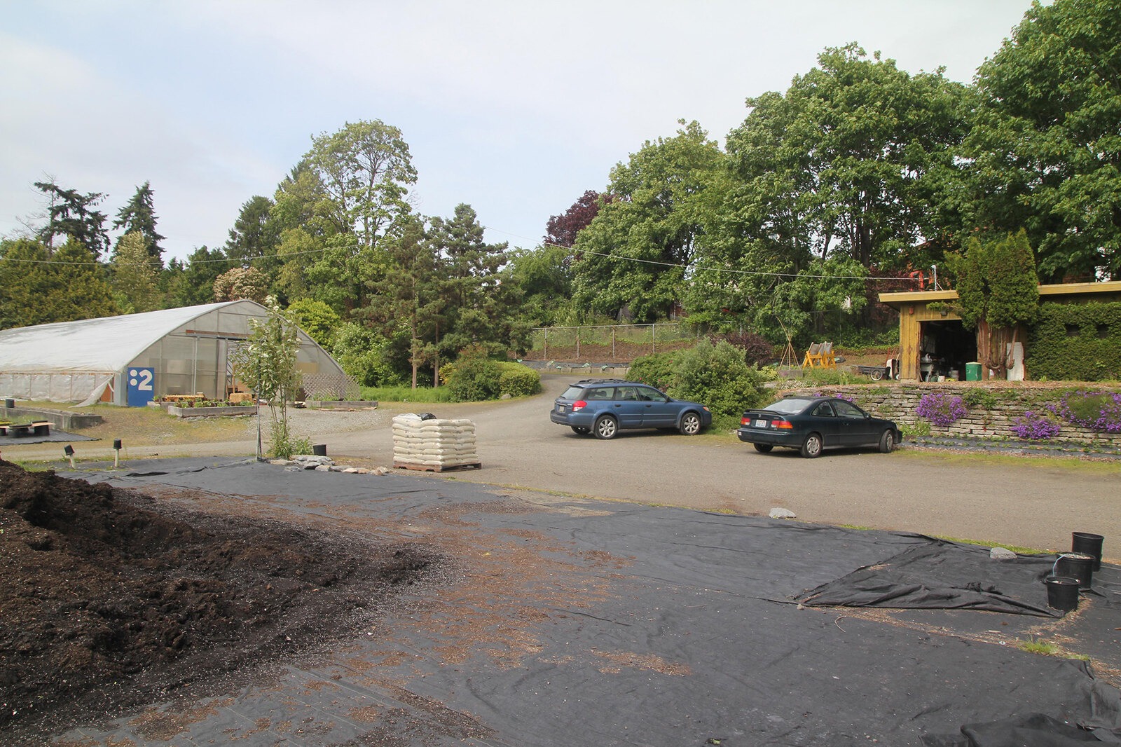 Gravel lot with parked cars, greenhouses and a small structure, showing the site before redevelopment with minimal landscaping.