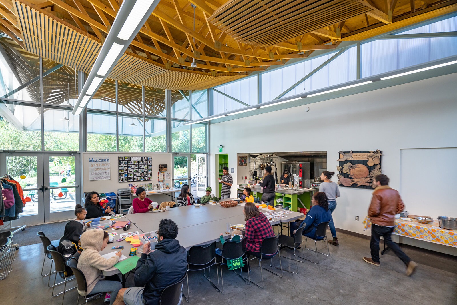 People of all ages gather around tables inside a bright community building with exposed wood beams, sharing food, crafts and conversation.