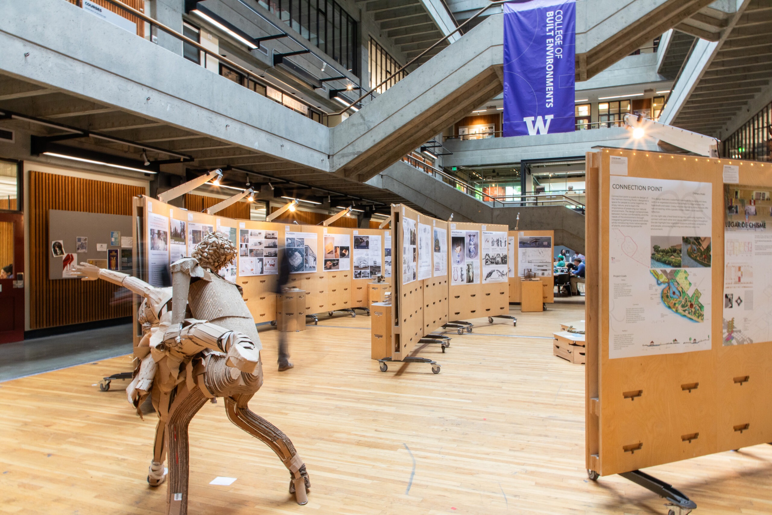Wide view of the Gould Hall atrium during the FrameWorks exhibition, featuring student design boards arranged in a circular layout, a large cardboard sculpture in the foreground and a College of Built Environments banner hanging above the space.