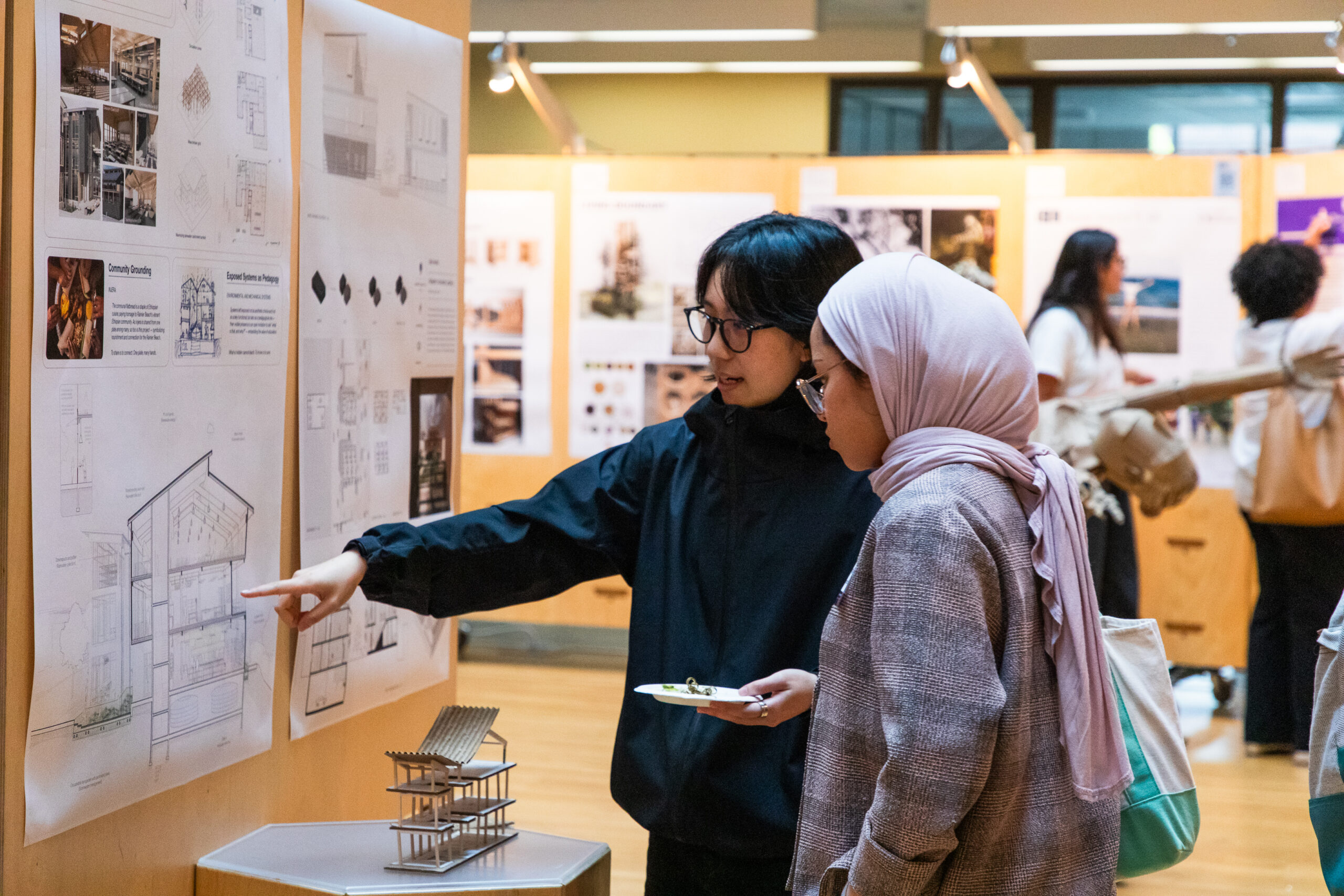 Two people stand beside an architectural presentation board, pointing at drawings and discussing a building section model displayed on a pedestal.