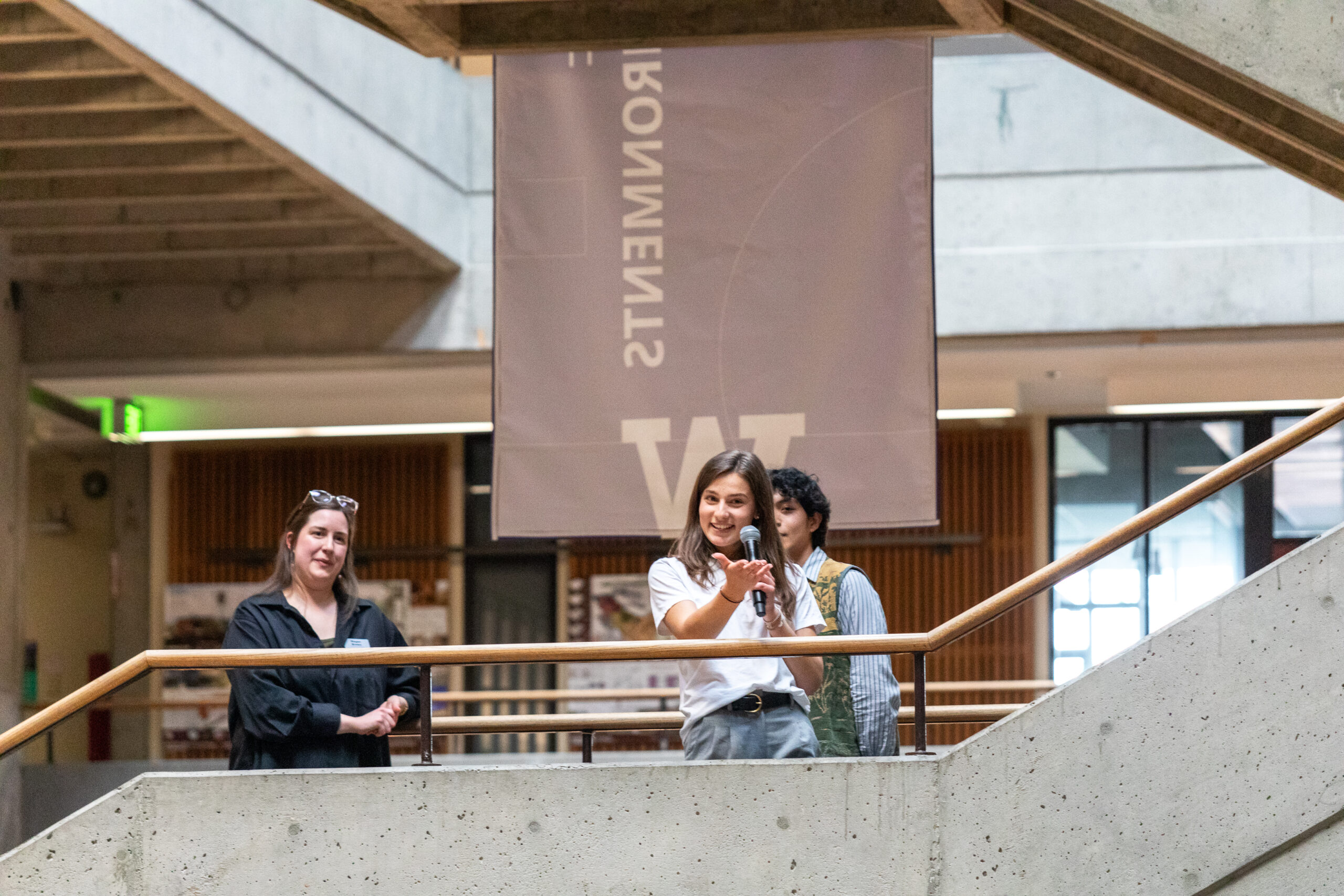 Three people speak from an upper-floor balcony in Gould Hall during the opening remarks for the FrameWorks exhibition.