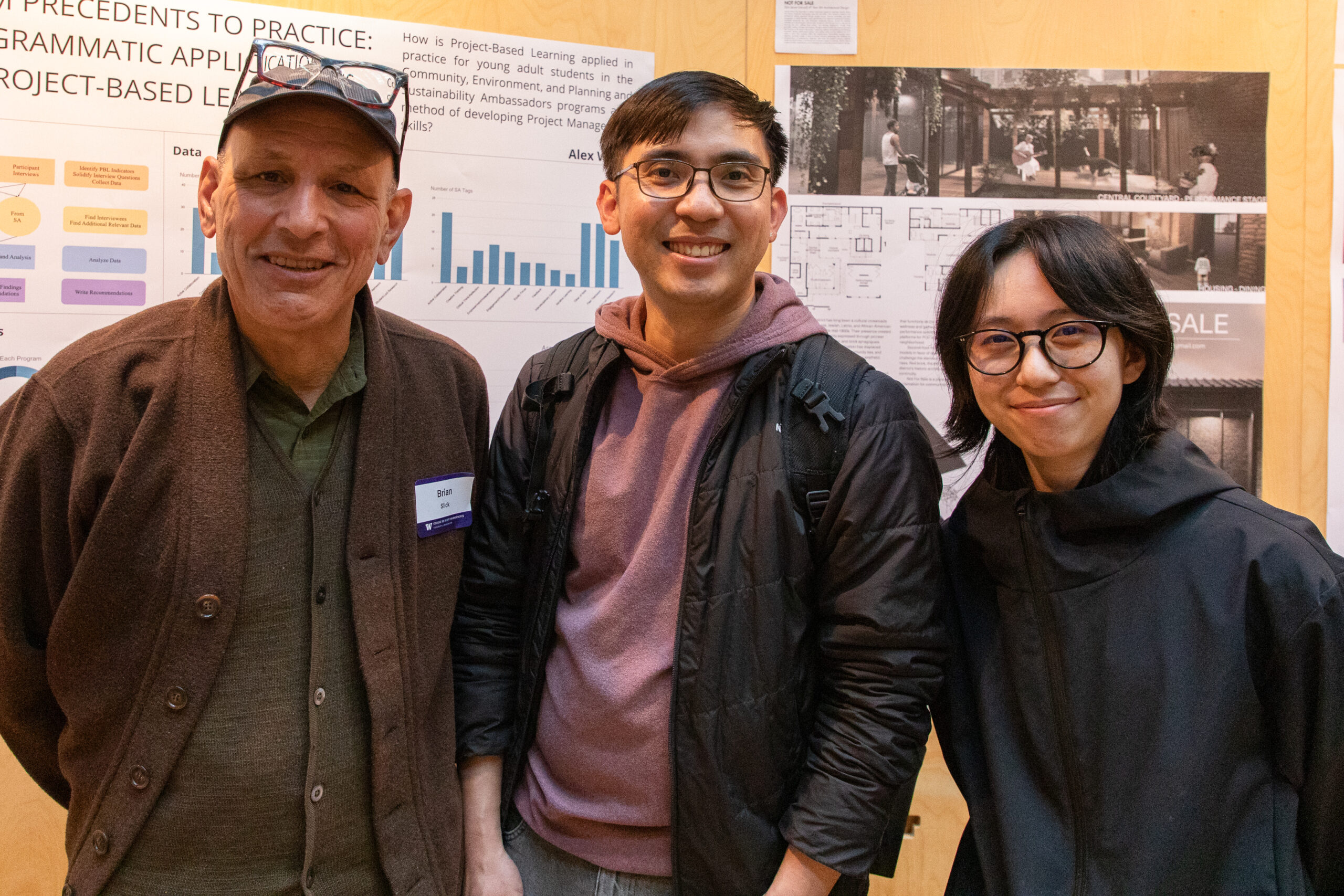 Three attendees pose in front of a student research and design presentation board during the exhibition reception.