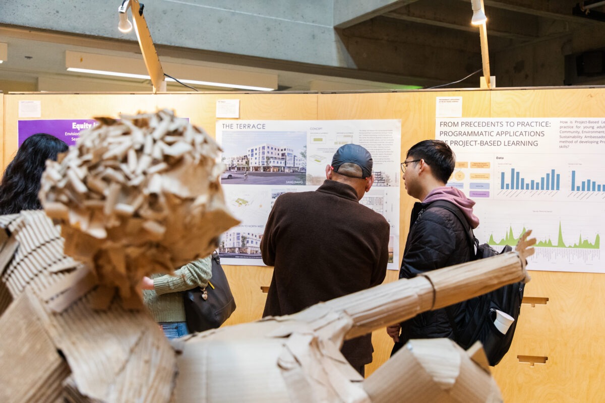 A cardboard human figure sculpture appears in the foreground while visitors review student work mounted on exhibition panels in the background.