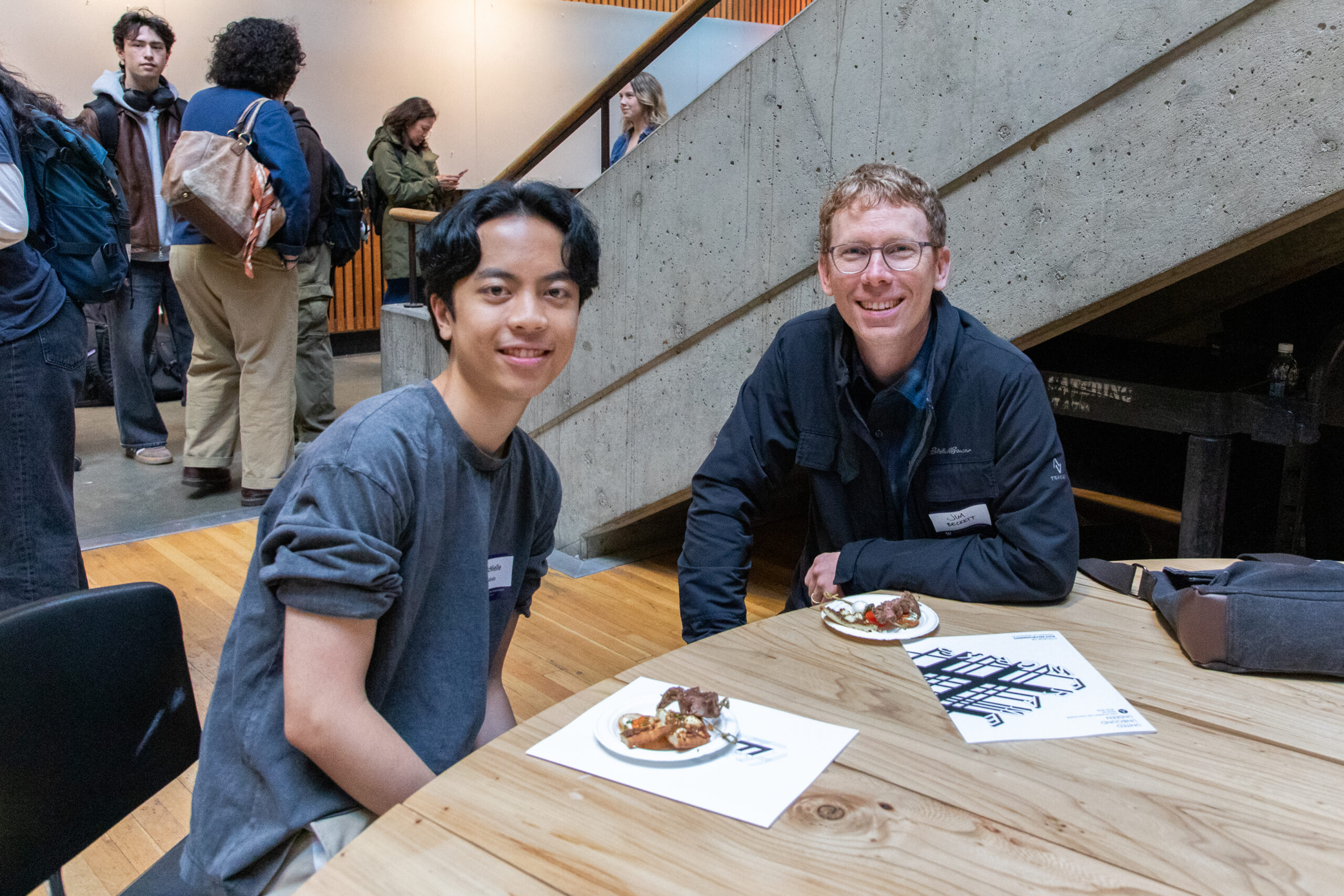 Two attendees seated at a table with appetizers and exhibition programs during the FrameWorks reception in Gould Hall.