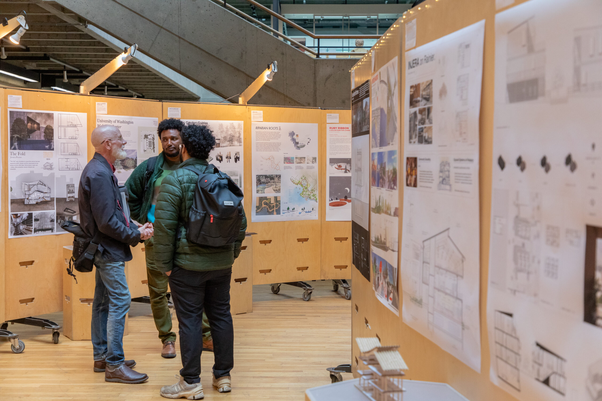 Visitors walk through the exhibition space while viewing student architecture and urban design projects displayed on wood panels.