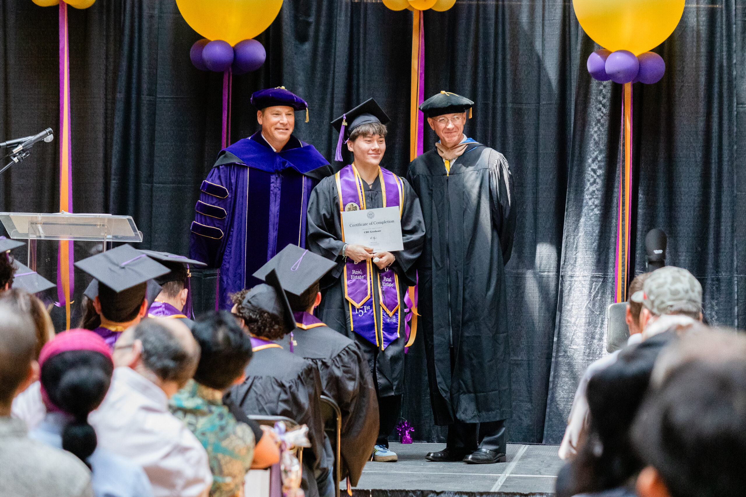 Student holding diploma on graduation stage with Dean and Chair