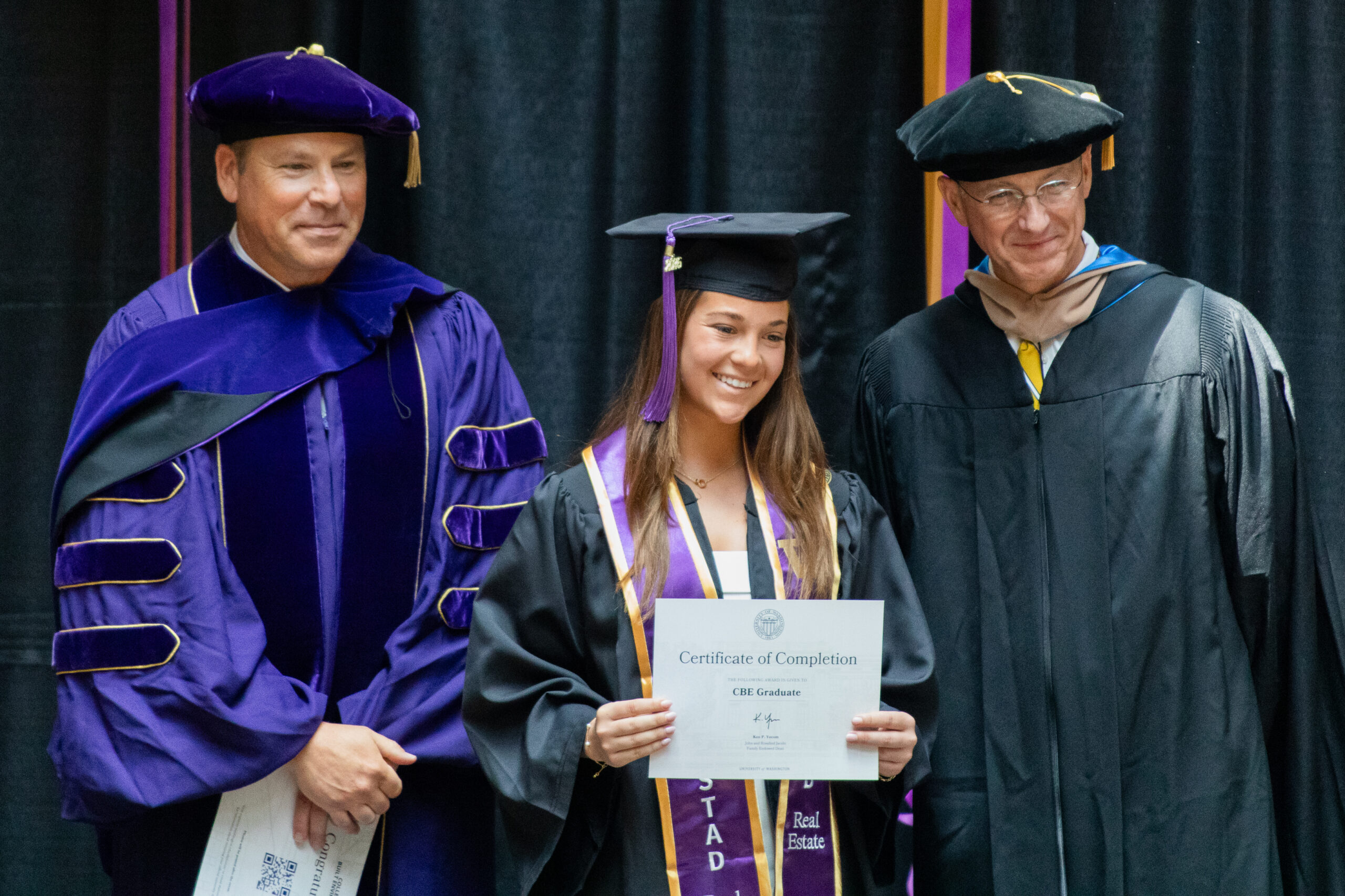Student holding diploma on graduation stage with Dean and Chair